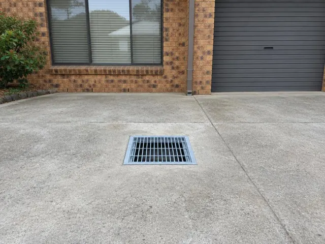 Square outdoor drain grate in a concrete driveway beside a brick house and garage door in Kings Langley